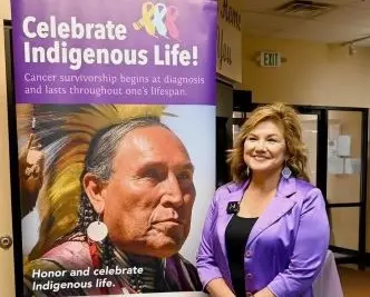 Woman dressed in purple standing next to banner entitled "Celebrate Indigenous Life"